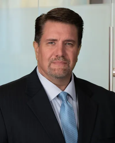 Middle-aged man in business suit and blue tie posing confidently against a glass office backdrop.