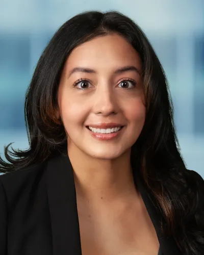 Smiling young woman with dark hair wearing a black blazer against a soft blue blurred background.