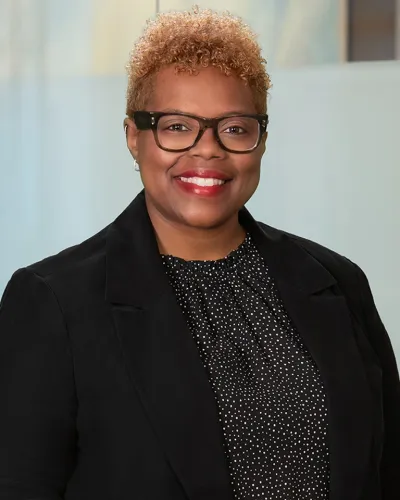 Smiling woman with curly hair wearing glasses, black blazer, and polka dot blouse in office setting.