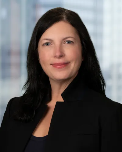 A smiling woman with long dark hair, wearing a floral shirt and dark cardigan against a neutral background.