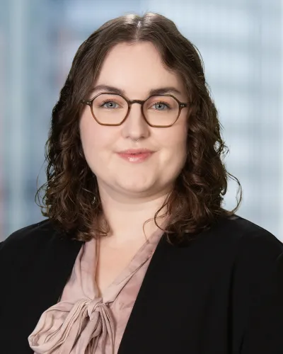 Smiling woman with curly brown hair and glasses wearing a pink tie-neck blouse against a neutral background