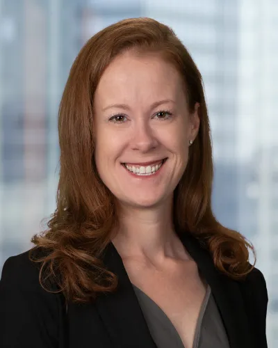Smiling woman with red hair wearing a black blazer and gray top in front of a blurred office building background.