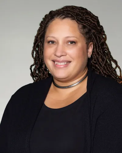Smiling woman with long braided hair wearing a black outfit and silver choker against a neutral background