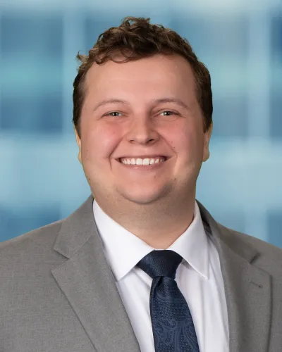 Smiling man in gray suit, white shirt, and patterned navy tie against a blurred blue background.