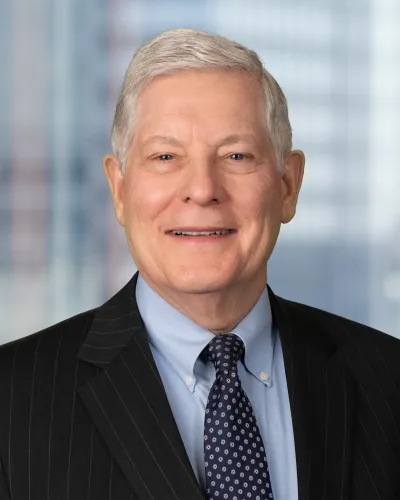 Older man in a dark pinstripe suit, blue shirt, and patterned tie smiling against blurred office background