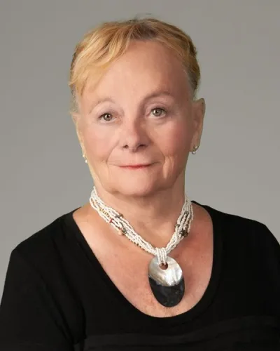 Portrait of an elderly woman with short blonde hair wearing a black top and a large decorative necklace.