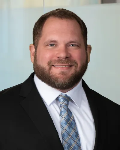 Smiling bearded man wearing a black sweater with gray hood and white shirt, posing against plain gray background