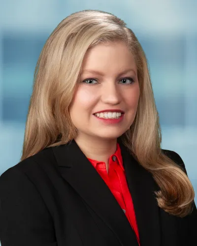 Professional woman with long blonde hair wearing a black blazer and red blouse smiling against blue background