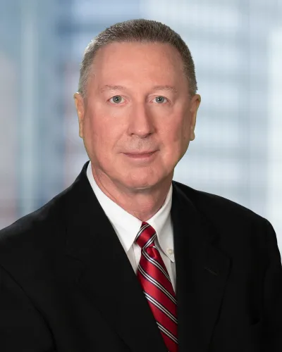 Professional headshot of a middle-aged man in a black suit and red striped tie against blurred office background