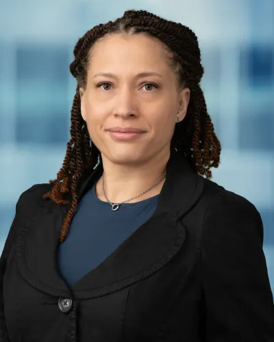 Professional woman with braided hair wearing black blazer and blue top against blurred office background