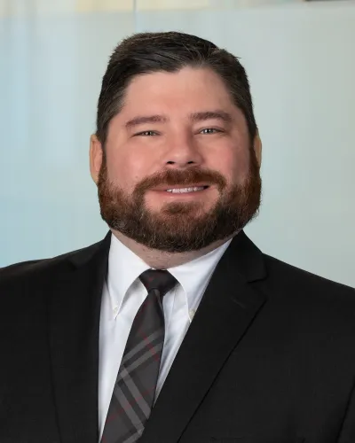 Smiling man with dark hair and beard wearing a houndstooth blazer and white shirt against gray background
