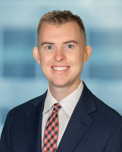 Professional headshot of a smiling young man in a navy suit and checkered tie with a blurred office background