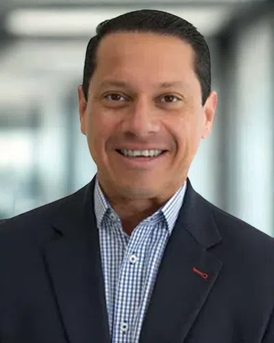Smiling businessman wearing a checkered shirt and dark blazer in a modern office setting.