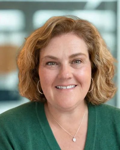 Smiling woman with curly light brown hair wearing a green V-neck sweater and silver necklace indoors.