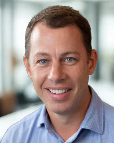 Smiling man with blue eyes and light brown hair wearing a blue shirt in a bright modern office setting.