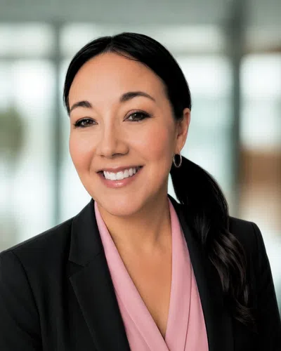 Smiling professional woman with dark hair in ponytail wearing black blazer and pink blouse in bright office.