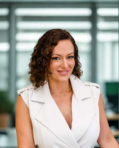 Smiling woman with curly hair wearing a white sleeveless blazer and brown belt in an office setting