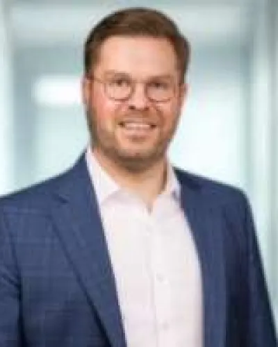 Smiling man with glasses wearing blue blazer and white shirt in a bright office setting.
