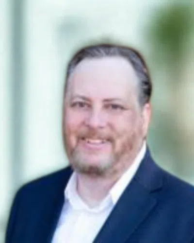 Middle-aged man with beard wearing a navy suit and white shirt smiling outdoors with blurred background