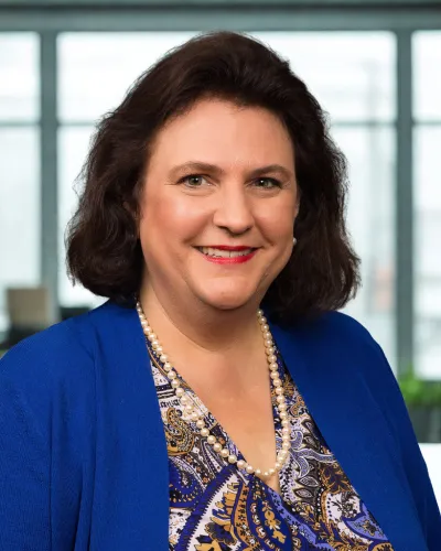 Smiling woman with dark hair wearing a blue cardigan, patterned blouse, and pearl necklace in office setting
