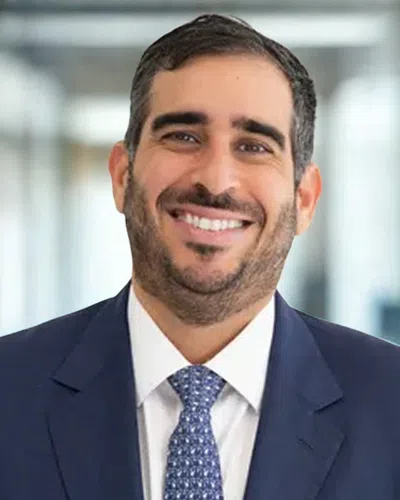 Smiling man in a navy blue suit and patterned tie with a blurred office background.