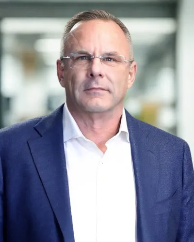 Professional middle-aged man wearing glasses, a navy blazer, and white shirt against a gray backdrop.