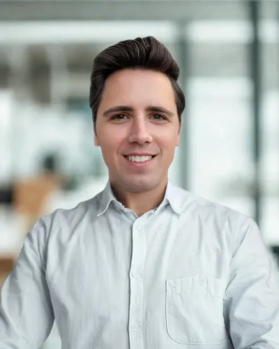 Young man with dark hair wearing a white shirt smiling against a gray textured background.