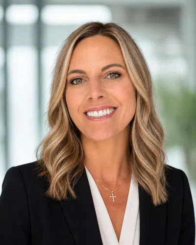 Professional woman smiling wearing a black blazer, white blouse, and a gold cross necklace with wavy blonde hair.