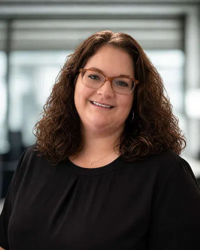 Smiling woman with curly hair and glasses wearing a black top in a bright modern office setting