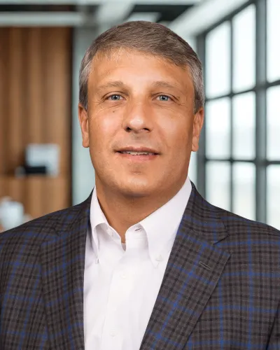 Middle-aged man in a plaid blazer and white shirt posing in a modern office setting with large windows.