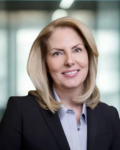 Professional woman with blonde hair smiling, wearing a black blazer and light blue blouse against gray background
