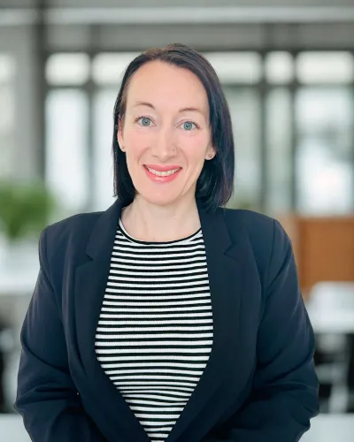 Smiling woman with black hair in a black blazer and striped shirt in a bright modern office setting.