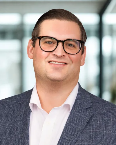 Smiling man with glasses wearing a gray suit jacket and white shirt standing in a modern office environment