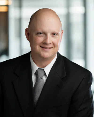 Smiling bald man in a black suit with gray tie posing in an office with blurred windows background