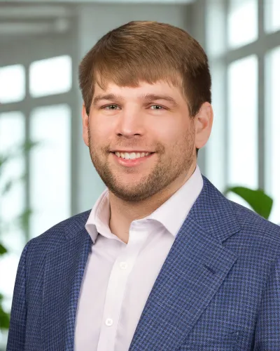 Smiling man with light brown hair wearing a blue checkered blazer and white shirt in a bright office setting