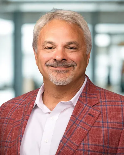 Smiling middle-aged man with gray hair and goatee wearing a red plaid blazer and white shirt in office setting