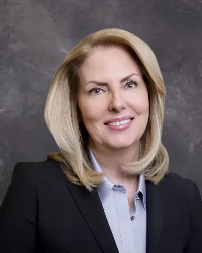 Professional woman with blonde hair smiling, wearing a black blazer and light blue blouse against gray background