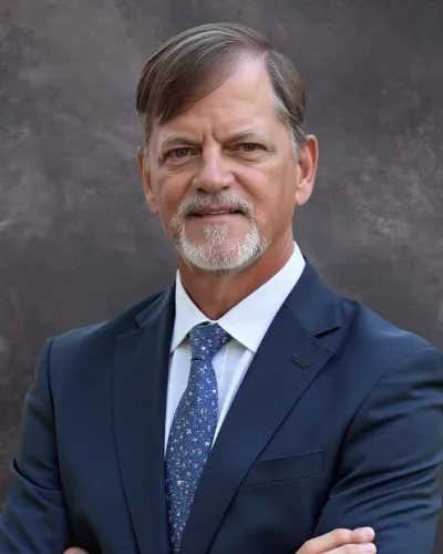 Middle-aged man with gray beard wearing navy suit and patterned blue tie against gray backdrop.