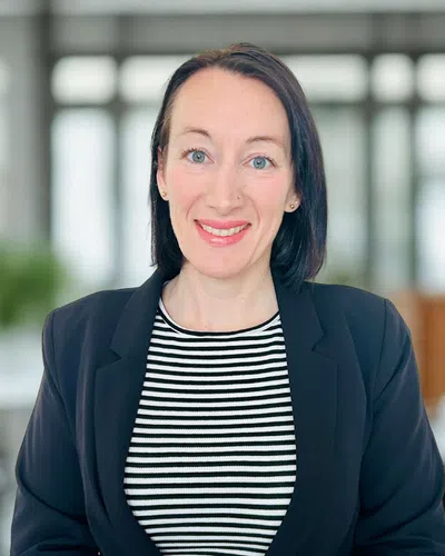Smiling woman with black hair in a black blazer and striped shirt in a bright modern office setting.