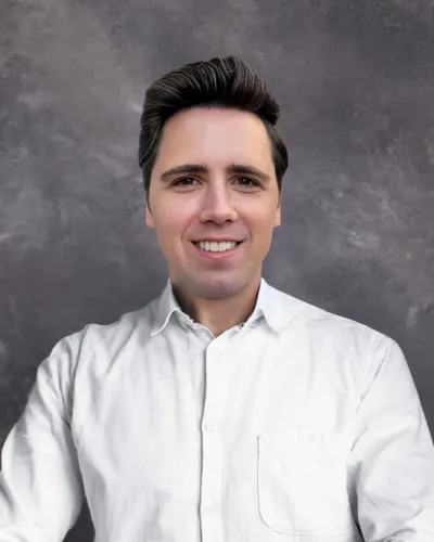 Young man with dark hair wearing a white shirt smiling against a gray textured background.
