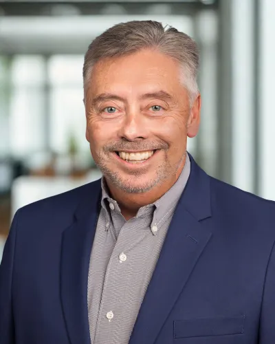 Smiling middle-aged man with gray hair wearing a blue blazer and gray checkered shirt in office setting