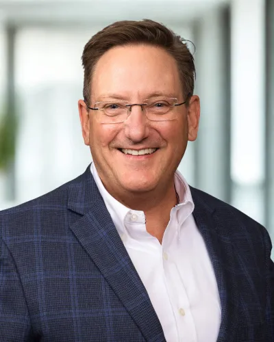Smiling middle-aged man wearing glasses, a blue checkered blazer, and a white shirt in a bright office setting.