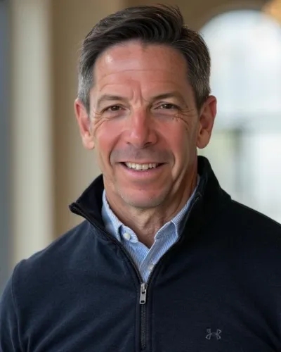 Smiling middle-aged man wearing a navy zip-up sweater and light blue shirt in a softly lit indoor setting