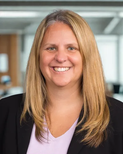 Smiling woman with long blonde hair wearing a black blazer and light purple top in an office setting.
