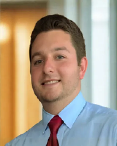Young professional man wearing light blue shirt and red tie smiling in office setting with blurred background.