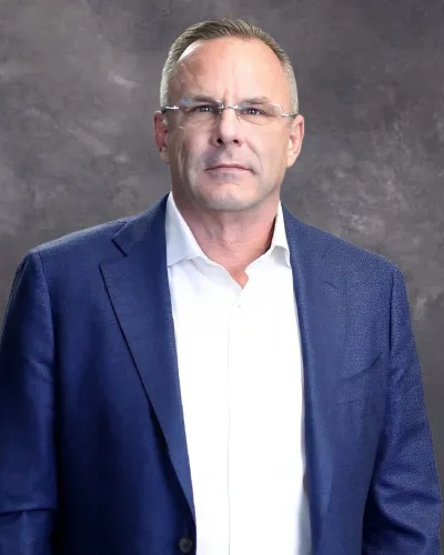 Professional middle-aged man wearing glasses, a navy blazer, and white shirt against a gray backdrop.