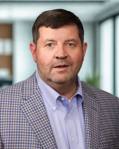 Middle-aged man wearing a checked blazer and light blue shirt posing in an office background.