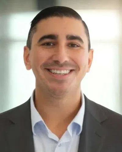 Smiling man in a suit jacket and light blue shirt standing in a bright, blurred office environment.