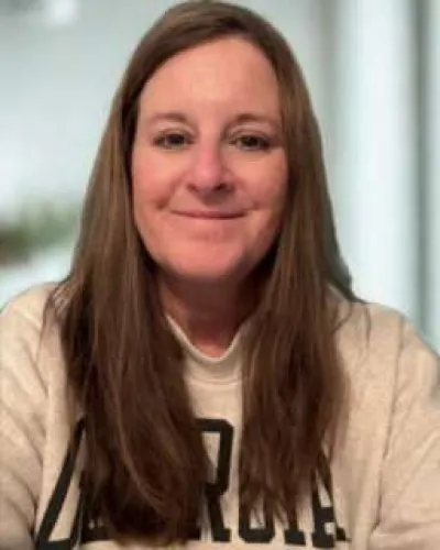 Smiling woman with long brown hair wearing a beige sweatshirt with bold black letters in a bright indoor setting