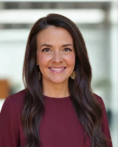 Smiling woman with long dark hair wearing burgundy top and gold earrings in a softly blurred indoor setting